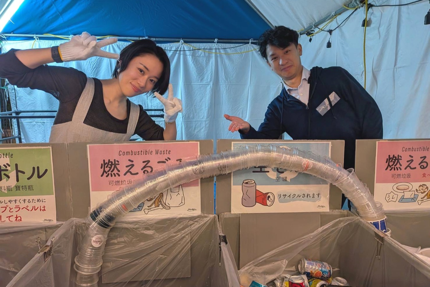 ごみ箱の上に架かった、重ねたプラスチックカップのアーチの後ろでポーズをとるUr AIのメンバー2人